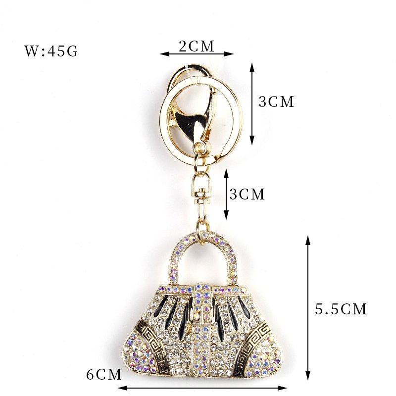 Rounded handbag-shaped pendant with arched handle, decorative strips, sparkling accents, ring and clip in cream, clear, gold-tone, shown as a keychain.