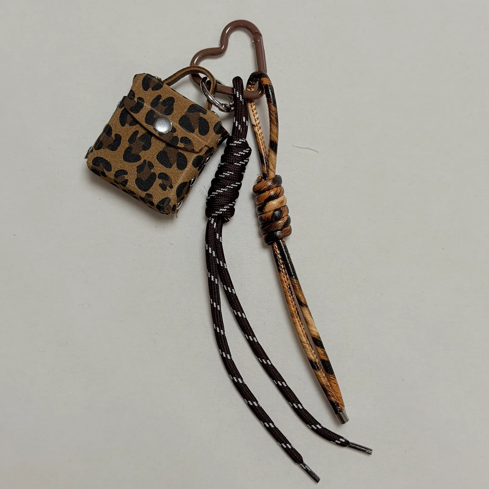Leopard-print miniature bag charm with knotted cord tails and a brown heart-shaped clasp.