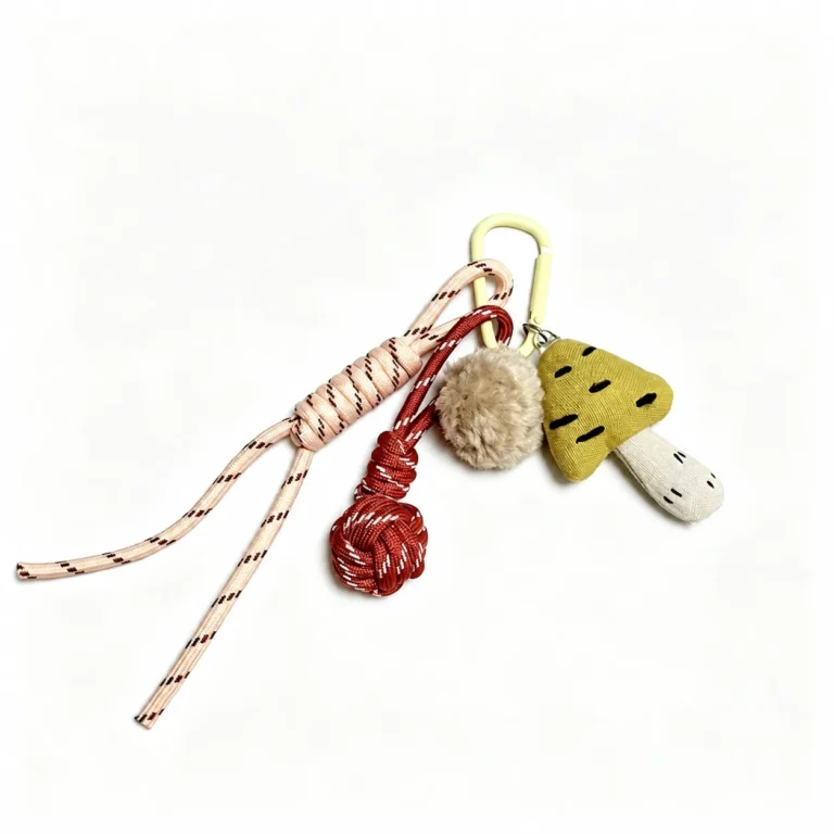 Fuzzy mushroom and pom-pom with cord tassels and clasp in cream, yellow, red, black, shown as a clip-on charm.
