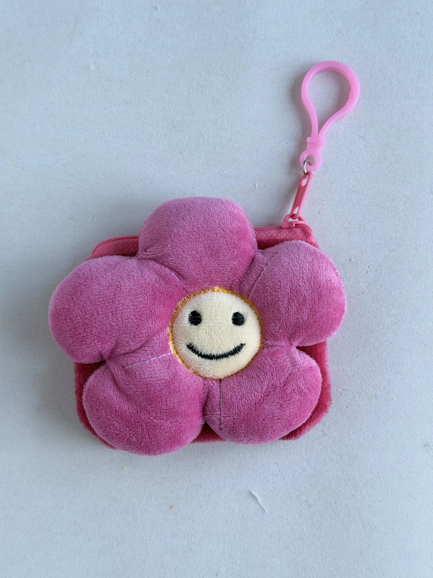 Pink flower-shaped fuzzy pouch with stitched-looking face details, a visible upper zipper and a pink clasp.