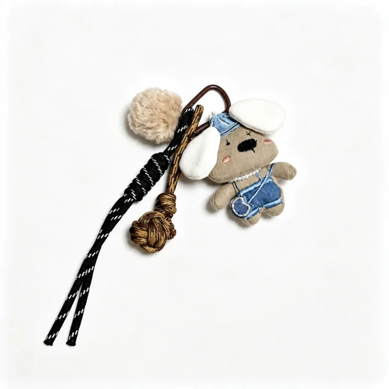Fuzzy long-eared dog with cap and outfit and pom-pom and clasp in tan, blue, white, brown, black, shown as a clip-on charm.