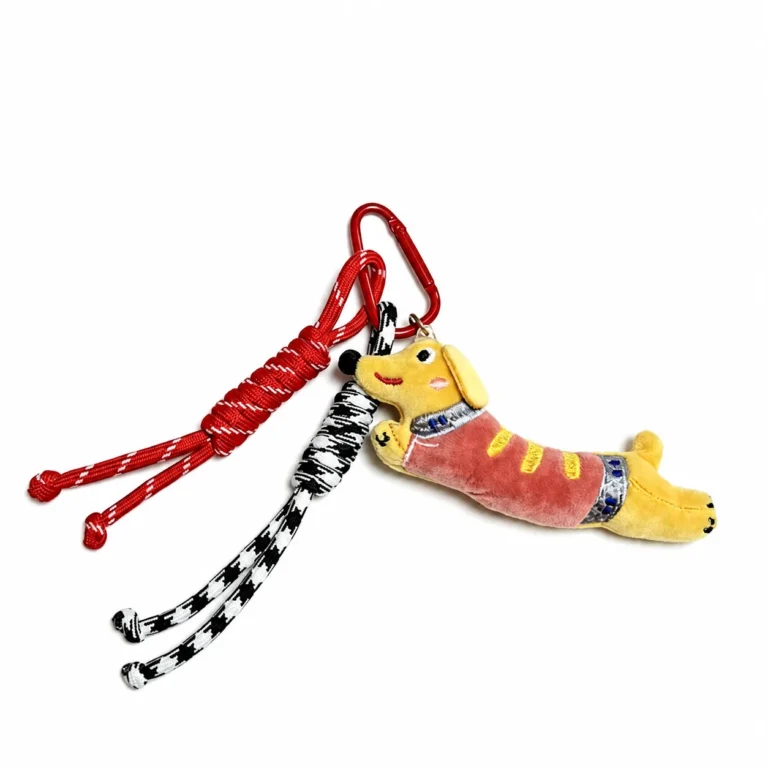 Fuzzy dachshund figure with cord tassels and triangular clasp in yellow, red, black, white, shown as a clip-on charm.